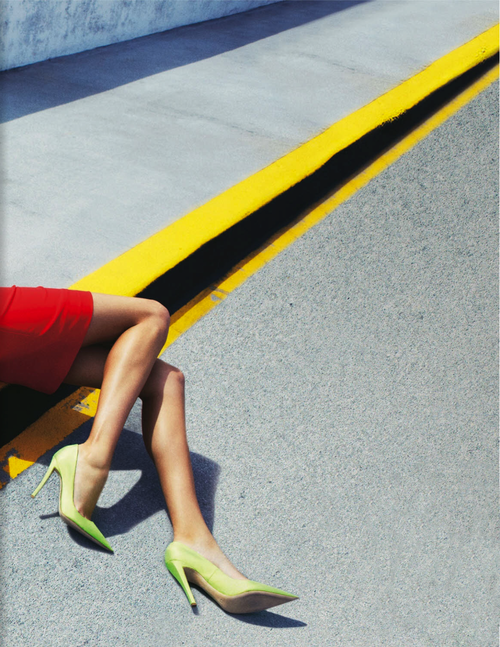 Femme en robe rouge allongé sur un trottoir avec des chaussures vertes. Une ligne jaune longe le trottoir