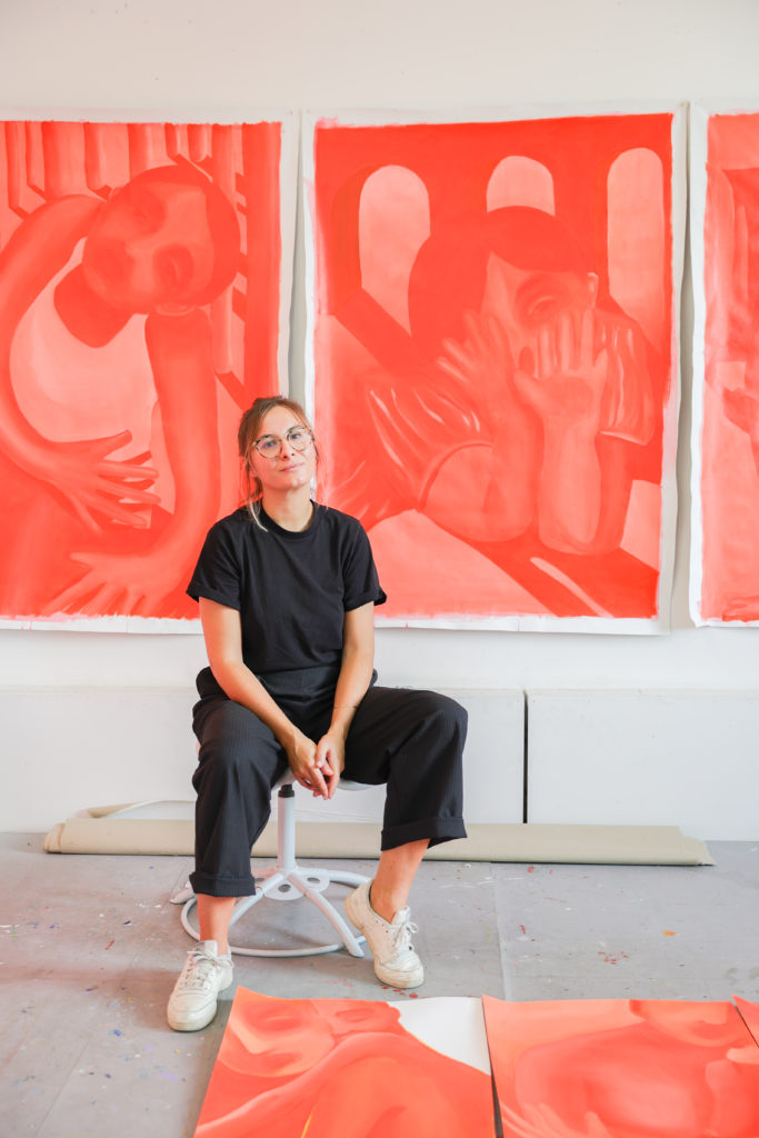 La peintre camille cottier habillé de noir assise sur un tabouret devant un triptyque de toile rouge