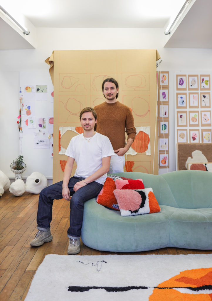 Le duo Amca Oval posant dans son studio de design. Les des deux hommes porte un tshirt blanc et un jean, est assis sur le canapé bleu. L'autre porte un haut marron et se tient debout derriere le canapé
